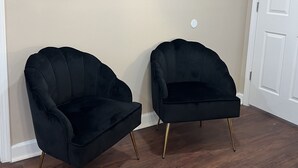 Una habitación con dos sillones negros de patas doradas, un espejo redondo en la pared y una puerta blanca.