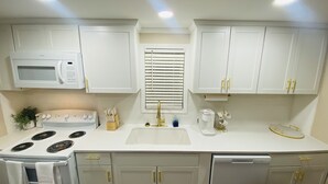 Una cocina con armarios blancos, microondas, estufa, fregadero y ventana con persianas.