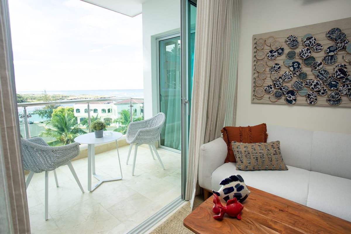 Een balkon met twee rieten stoelen, een klein rond tafeltje met een bloempot en een witte bank met decoratieve kussens.