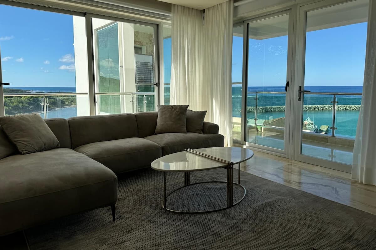 Een moderne woonkamer met een grote hoekbank, een ronde salontafel en uitzicht op de oceaan door middel van kamerhoge ramen.