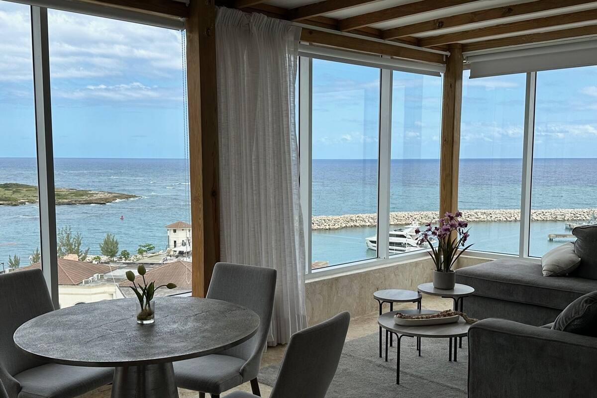 Een kamer met een groot raam met uitzicht op zee, een ronde tafel met stoelen en een bank.