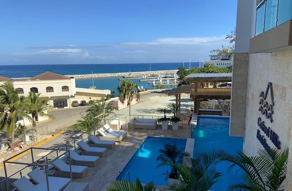 A pool area with lounge chairs, a view of the sea, and a building with a sign.