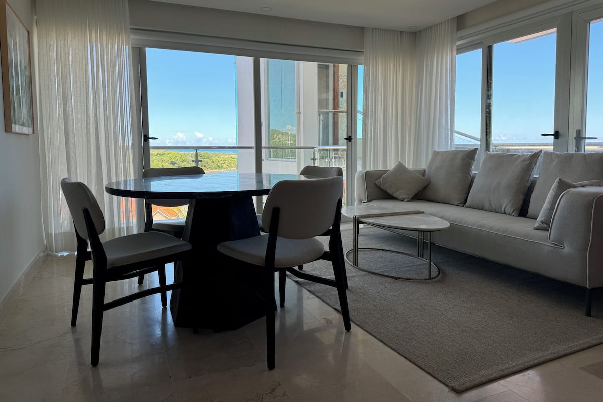 Een moderne woonkamer met een eettafel, stoelen, een bank en grote ramen met uitzicht op de zee.