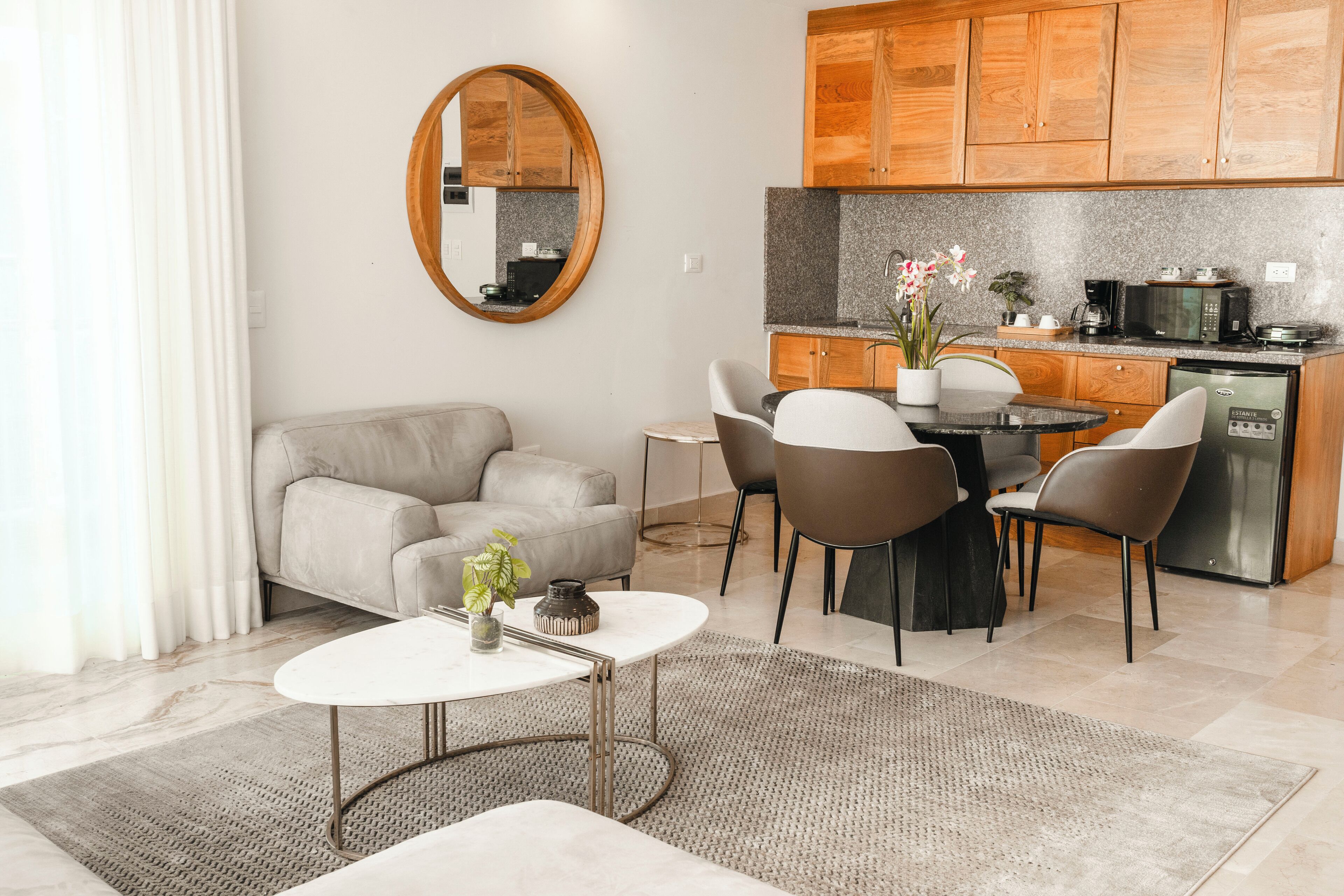 A modern living room with a sofa, coffee table, and dining area. A kitchen with wooden cabinets and appliances is visible in the background.
