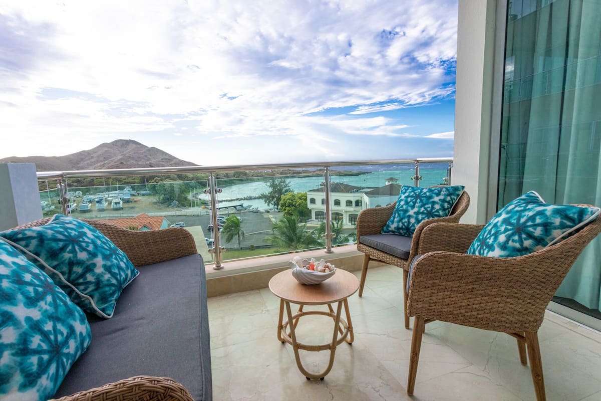Een balkon met rieten meubilair, een kleine ronde tafel en uitzicht op zee en gebouwen.