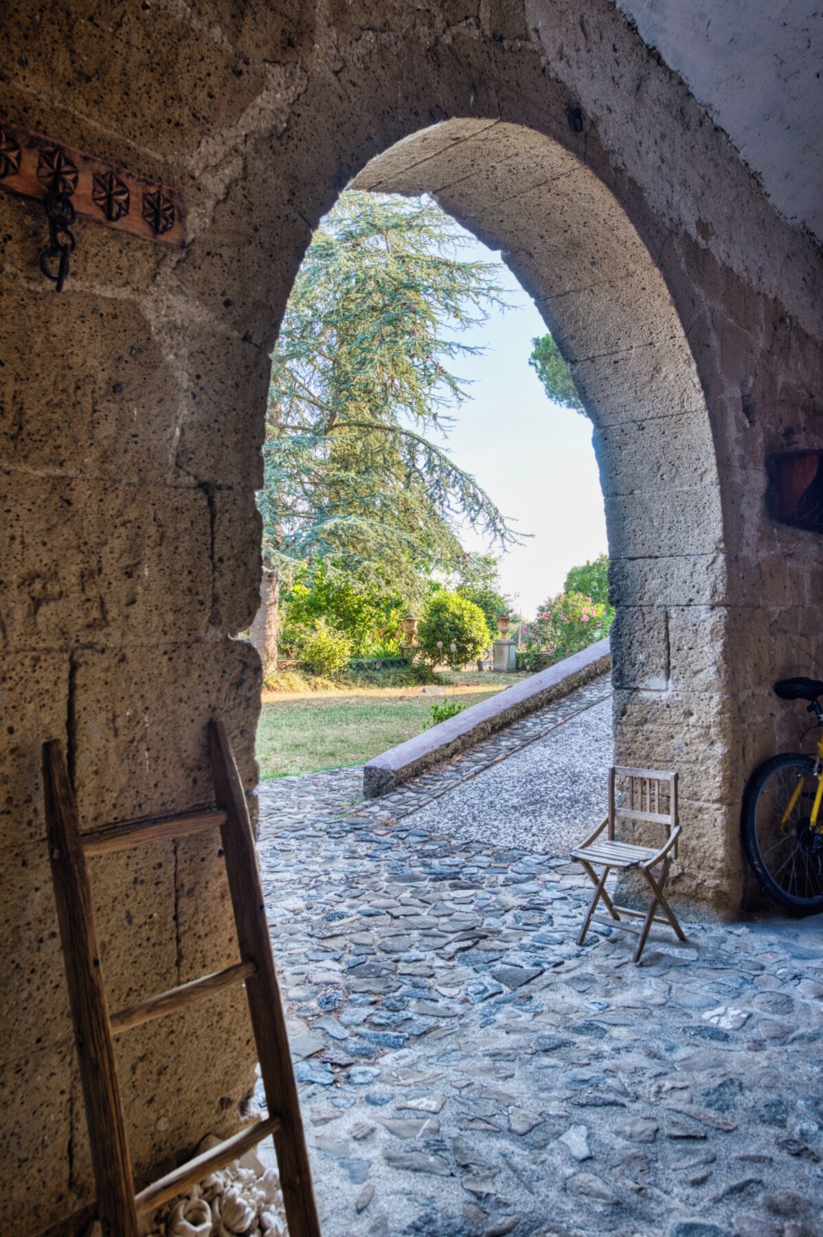 Una porta d'arc de pedra que dona a un camí de pedra i una cadira de fusta, a més d'una bicicleta.