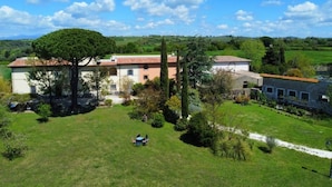A large, two-story building with a red-tiled roof, surrounded by a lush green lawn and trees. - The Borgo Renaissance Stable (Farnese)