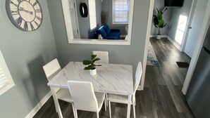 Een eetgedeelte met een marmeren tafel en witte stoelen, een grote wandklok, en een spiegel die een zithoek met een blauwe bank weerspiegelt.