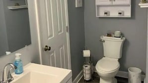 Een badkamer met een witte wasbak, spiegel en toilet. Er zijn planken met toiletartikelen en een wit kastje boven het toilet.
