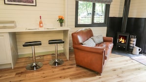 Une pièce chaleureuse avec un plancher en bois, un canapé en cuir, un coin bar avec des tabourets, une cheminée et une fenêtre donnant sur les arbres.