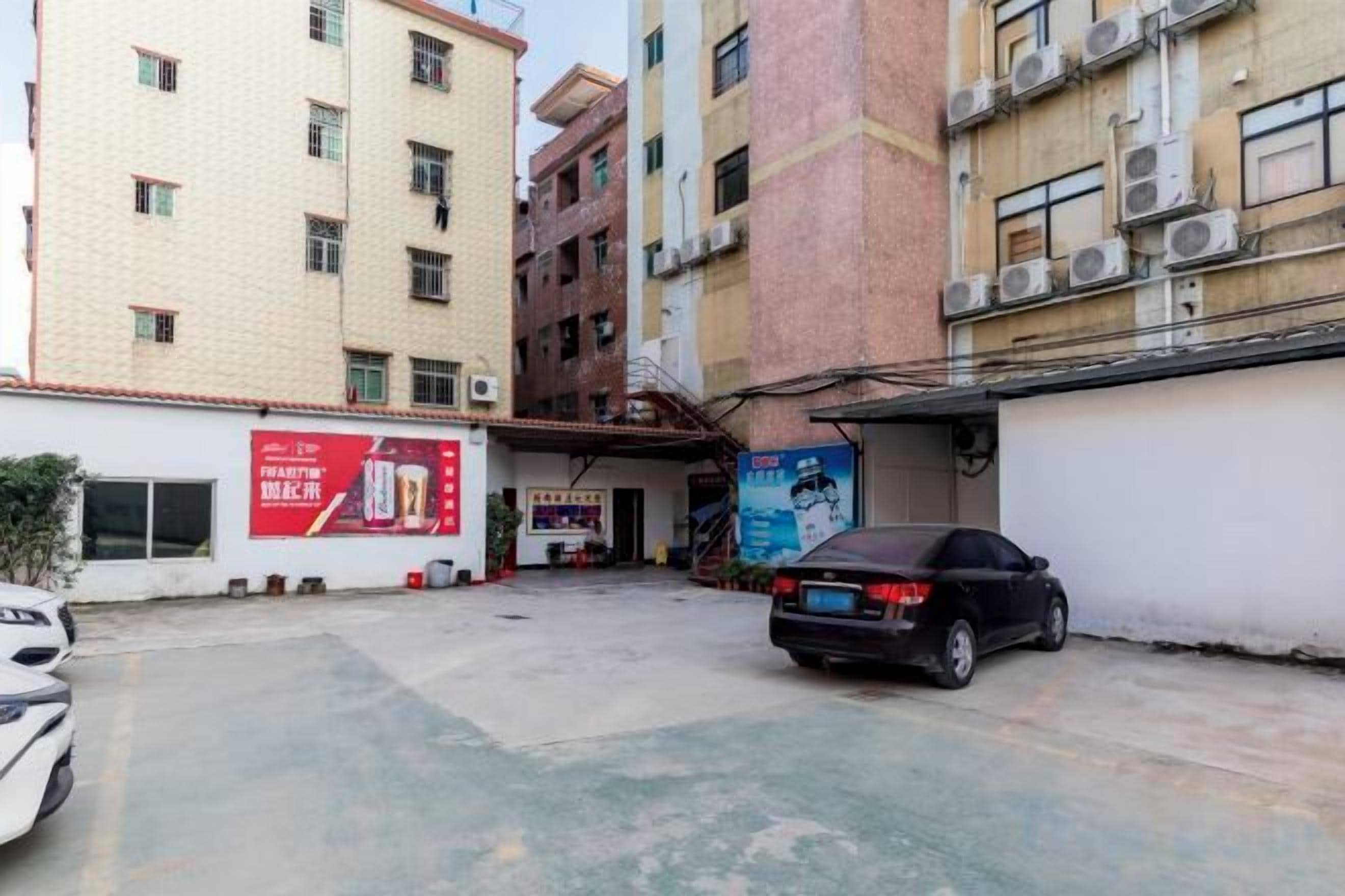 A parking area with a black car, a building with multiple floors, and a red advertisement on the wall.