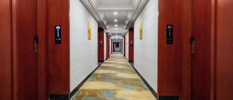 A long hallway with wooden doors on either side, carpeted with a wavy pattern, and a ceiling with recessed lighting.