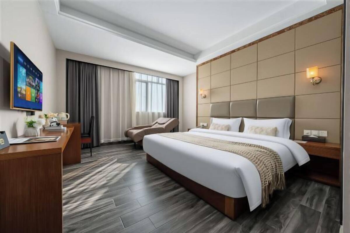 A hotel room with a large bed, a desk with a computer, a chair, a TV, and a window with curtains.