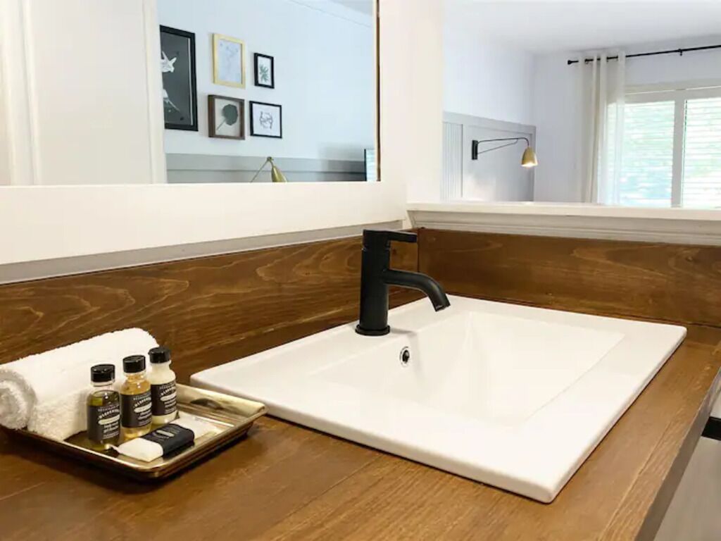 Un baño con lavamanos blanco, grifo negro, encimera de madera y una bandeja con artículos de aseo.