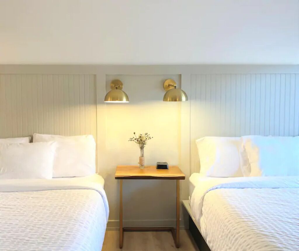 Habitación de hotel con dos camas, una mesita con un florero y dos lámparas de pared.