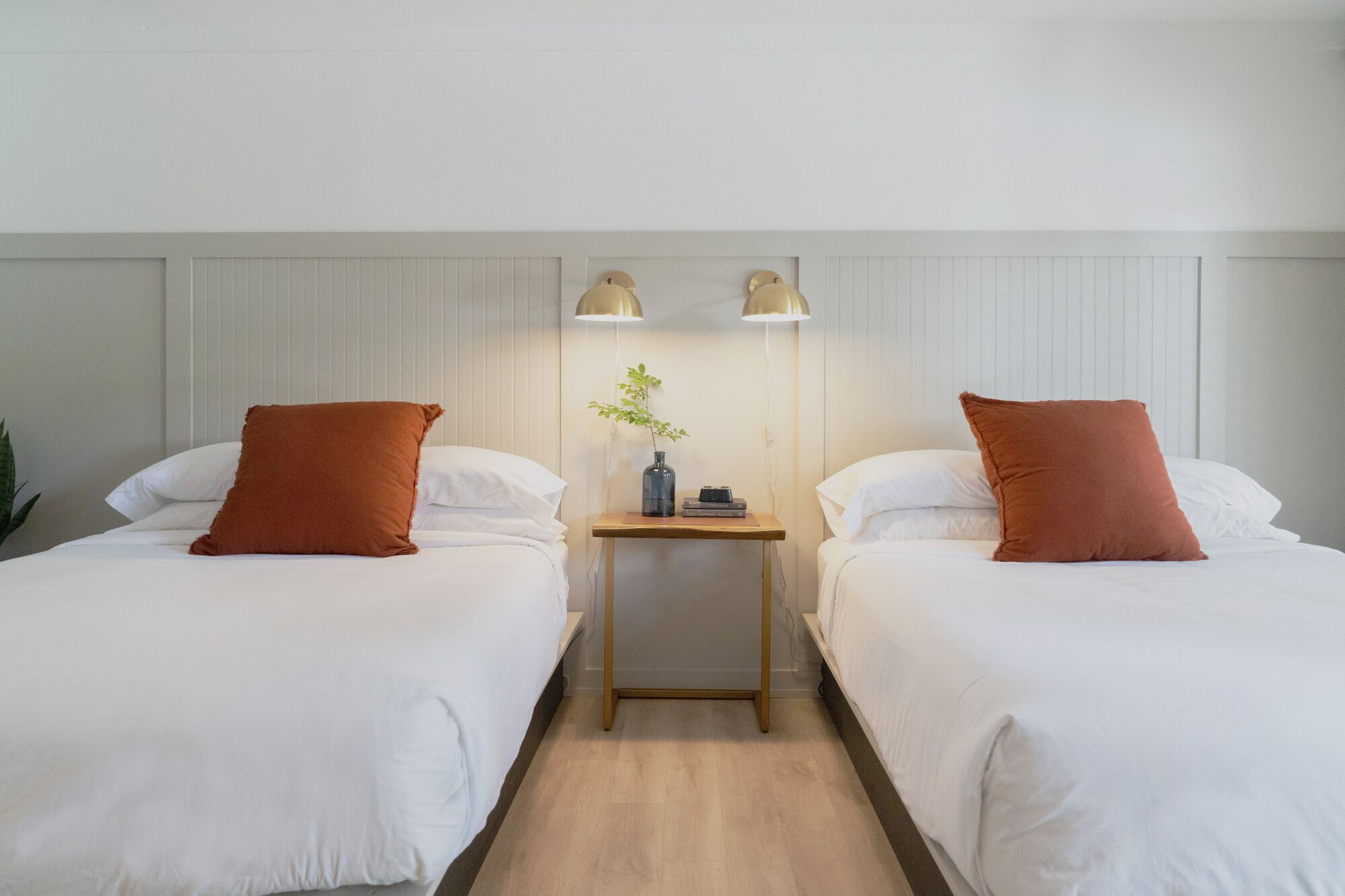 Habitación de hotel con dos camas, cada una con ropa de cama blanca y un cojín marrón decorativo. Entre las camas hay una mesita de noche de madera con una lámpara y un jarrón con una planta.