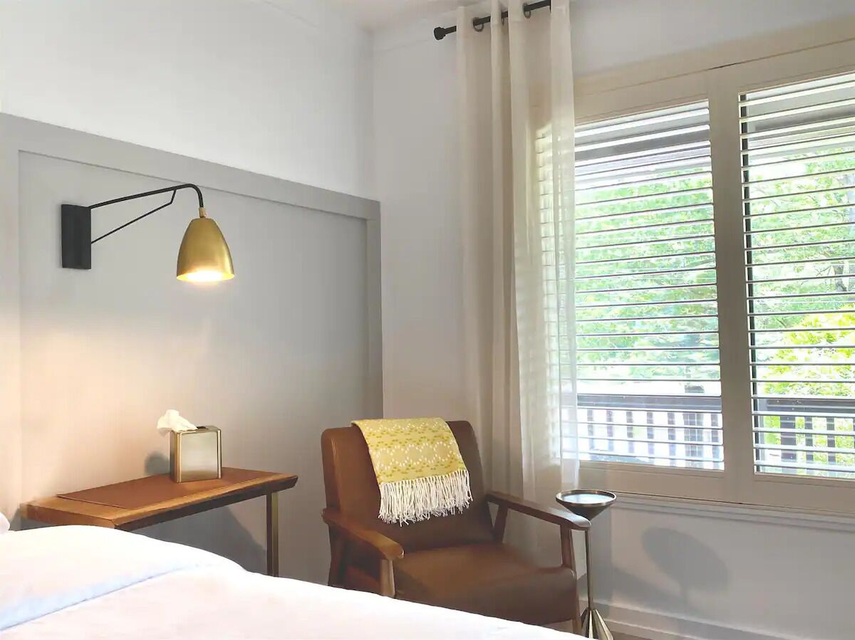 A hotel room with a bed, a chair with a yellow blanket, a bedside table with a lamp, and a window with blinds.