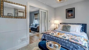 A bedroom with a large bed, a bedside table with a lamp, a mirror with a wooden frame, and a picture on the wall. - Forth House - Butterfly Getaway in Historic SPR! (Jacksonville)