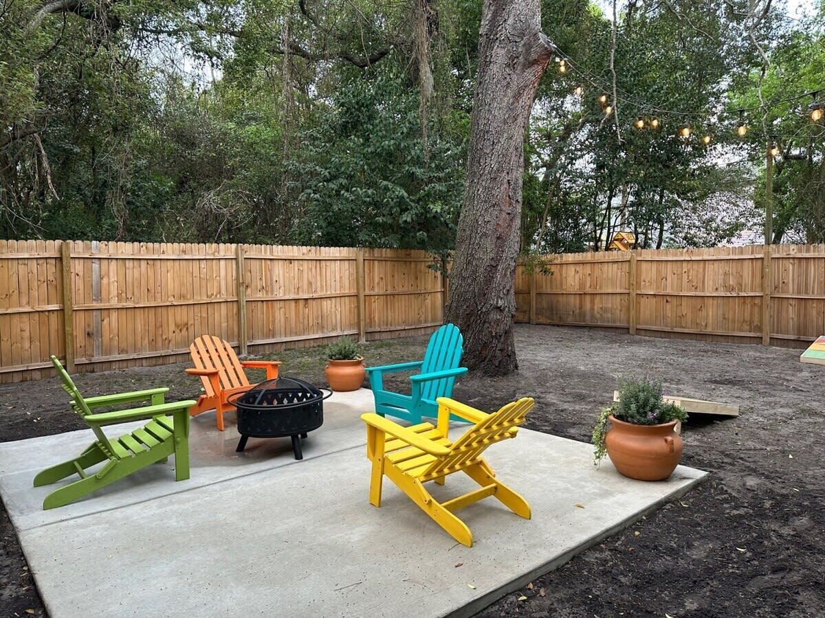 Un patio trasero con suelo de concreto, cerca de madera y mobiliario exterior que incluye sillas y un brasero.