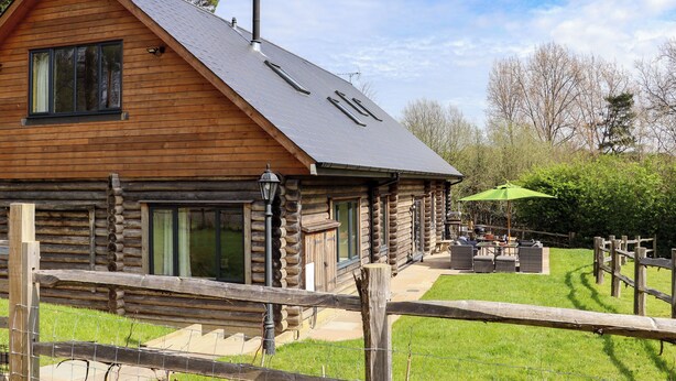 A log cabin with a large window, a chimney, and a patio area with outdoor furniture. - Ghyll Park Farm (Heathfield)