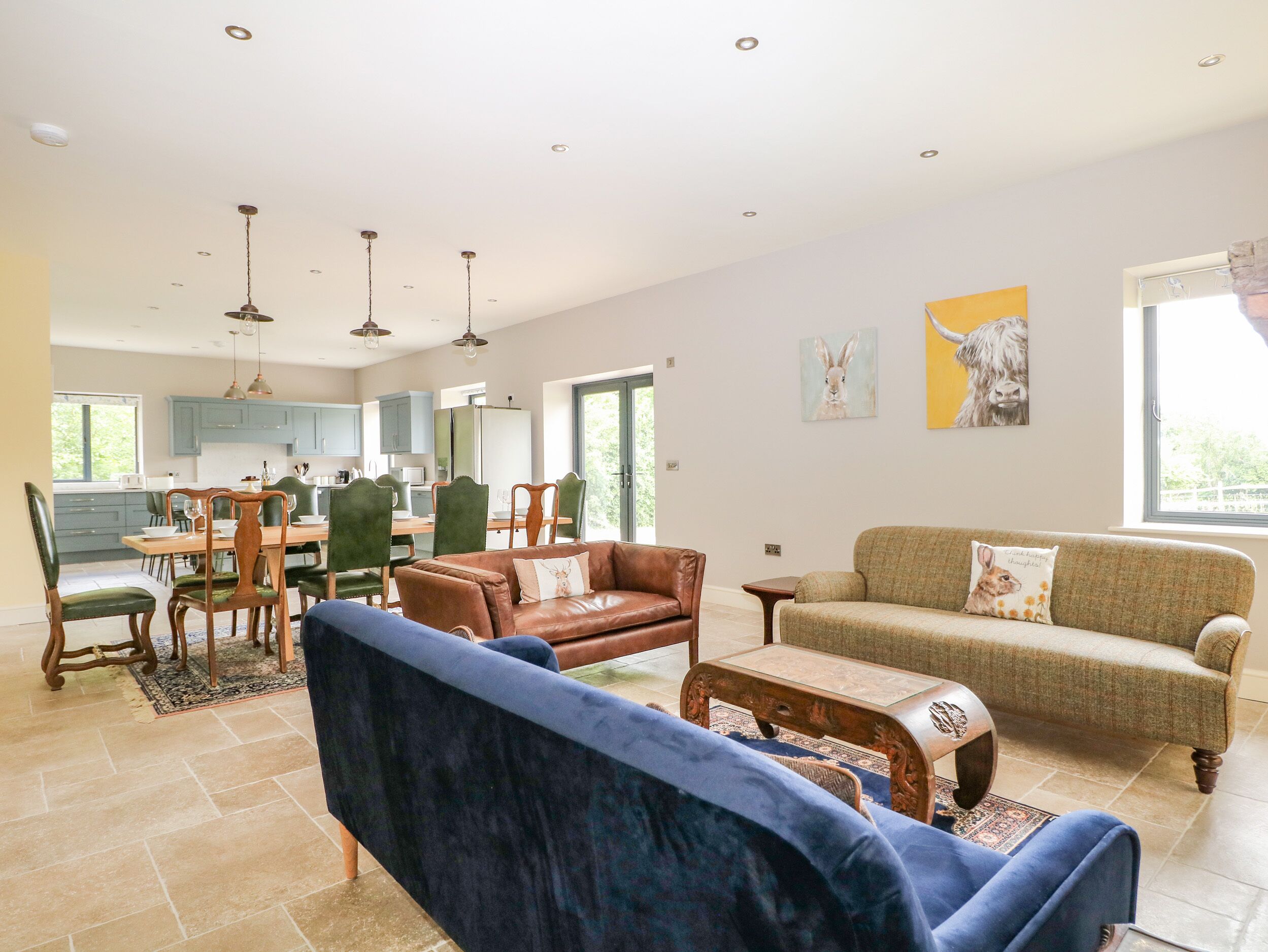 A spacious living room with a blue sofa, a brown leather couch, and a green sofa. There is a dining area with a wooden table and chairs. The kitchen is visible in the background with blue cabinets.