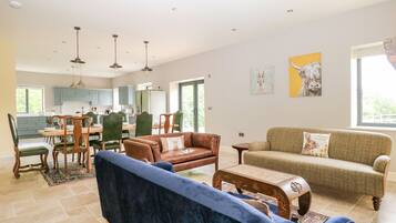 A spacious living room with a blue sofa, a brown leather couch, and a green sofa. There is a dining area with a wooden table and chairs. The kitchen is visible in the background with blue cabinets.
