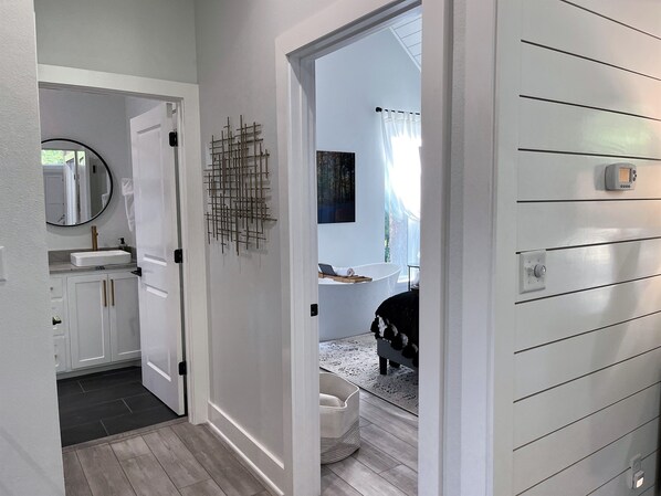 Un couloir mène à une salle de bain équipée d’un lavabo blanc, d’un miroir et d’un luminaire fixé au mur.