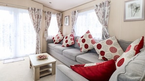 Ein modernes Wohnzimmer mit einem grauen Sofa, roten und weißen Kissen mit Muster sowie einem kleinen Holztisch und durchscheinenden Vorhängen mit floralem Dekor.