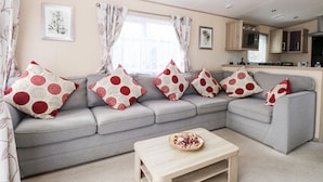 Ein Wohnzimmer mit grauem Sofa, roten und weißen Kissen mit Muster sowie einem Holztisch und einer Küche im Hintergrund.