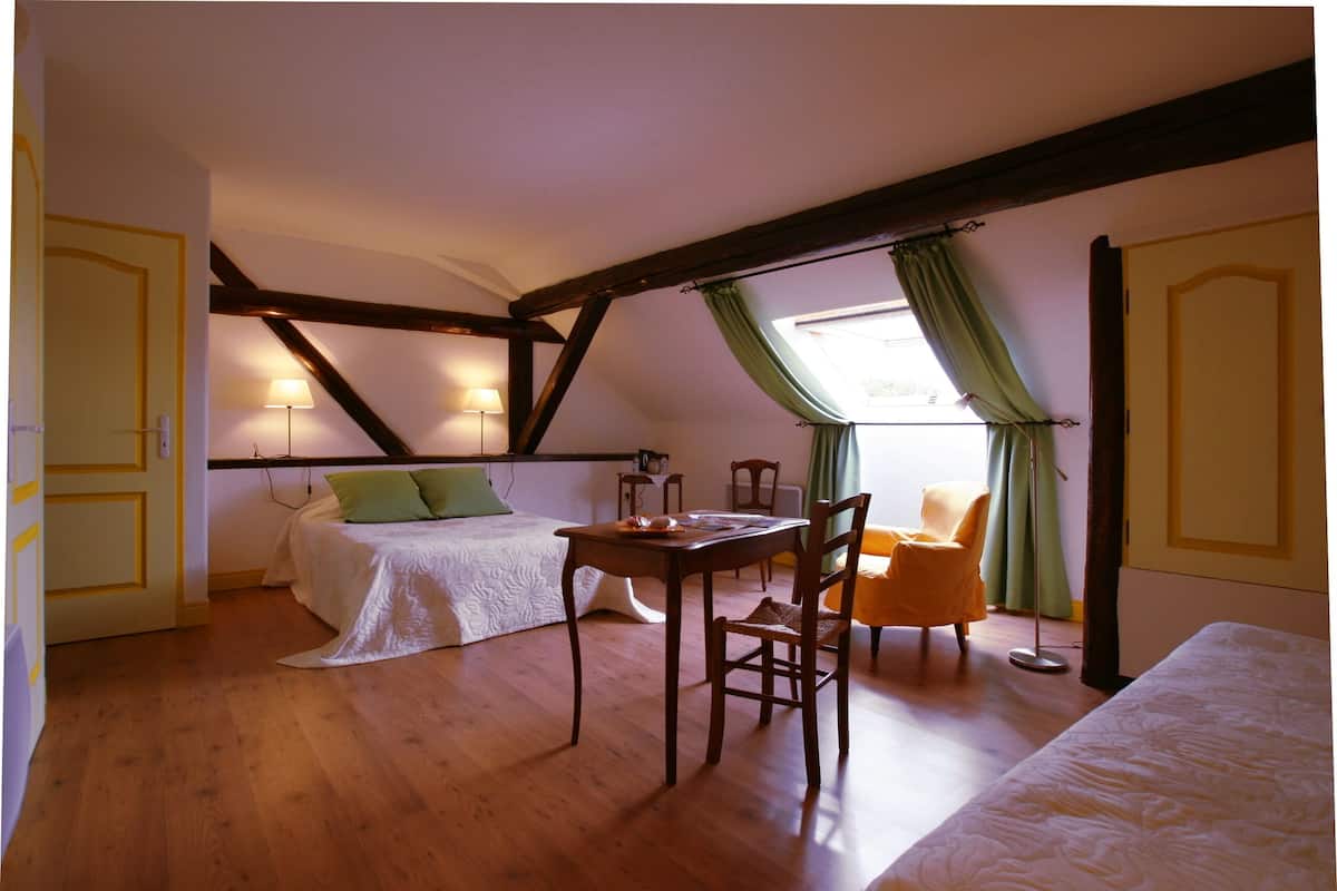 Une chambre avec un plancher en bois, un lit recouvert d’une courtepointe blanche, une table avec des chaises et une fenêtre avec des rideaux.