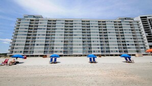 A large hotel with multiple floors and balconies. There are several beach umbrellas and lounge chairs on the sandy ground in front of the hotel. - Sands Beach Club II 625 - Ocean Front 2 Bed/2 Bath Condo - Amenities: Pools, Hot Tub & Tennis Court (Myrtle Beach)