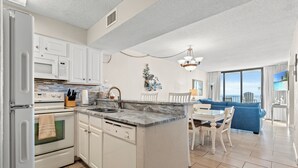 A modern kitchen with white cabinets, a built-in oven, and a dishwasher. There is a dining area with a blue sofa and a chandelier. - Sands Beach Club II 625 - Ocean Front 2 Bed/2 Bath Condo - Amenities: Pools, Hot Tub & Tennis Court (Myrtle Beach)