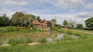 Perinteinen tiilitalo, jonka edessä on lampi ja jota ympäröi vehreä puutarha ja puut.