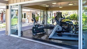 Un gimnasio al aire libre cubierto con equipo de ejercicios, incluyendo cintas de correr y bicicletas estáticas, y una puerta de vidrio que da a un patio.