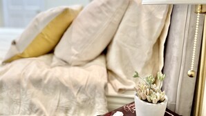 Una cama bien hecha con edredón blanco y beige, un cojín dorado y una mesita de noche con una pequeña planta en maceta.