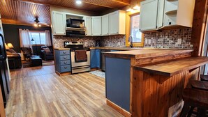 A spacious kitchen with wooden flooring, a central island, and a dining area with bar stools. - Pinetree Cabin - 4 king beds, stunning views (Custer)