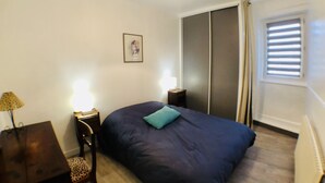 Une chambre à coucher comprenant un lit, une table de chevet, une lampe, une armoire et une fenêtre avec des stores.