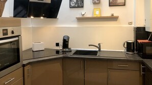 Une cuisine moderne équipée d’un four encastré, d’une machine à café, d’un évier et d’un grille-pain. Des tableaux encadrés ornent le mur, et une petite étagère présente un objet décoratif.