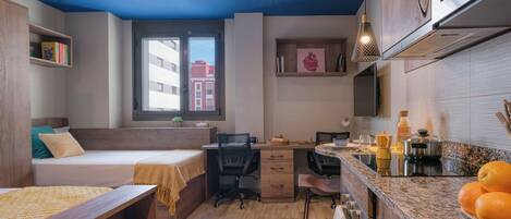 A compact studio apartment with a bed, desk, kitchenette, and a large window providing natural light.