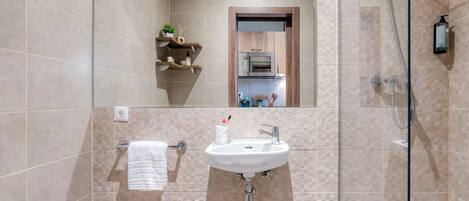 Un baño moderno con ducha de mampara de vidrio, un lavamanos blanco y un estante de madera con artículos de aseo.