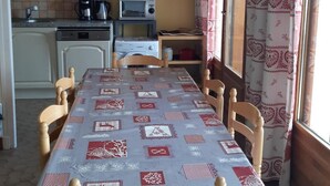 Ein Essbereich mit Tisch, bedeckt mit einer gemusterten Tischdecke, Holzsstühlen, einer Küche mit weißen Schränken, einer Mikrowelle und einer Kaffeemaschine.