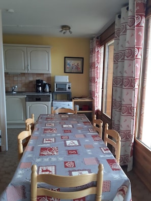 A dining area with a table covered in a patterned tablecloth, wooden chairs, a kitchen with white cabinets, a microwave, and a coffee maker.