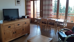 Ein Zimmer mit Holzschrank, Fernseher, Esstisch mit Stühlen und einer Couch.