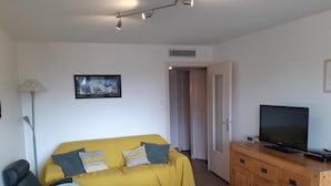 Ein Wohnzimmer mit gelbem Sofa, einem Holzschrank mit Fernseher, einem Sessel und einem kleinen Tisch.