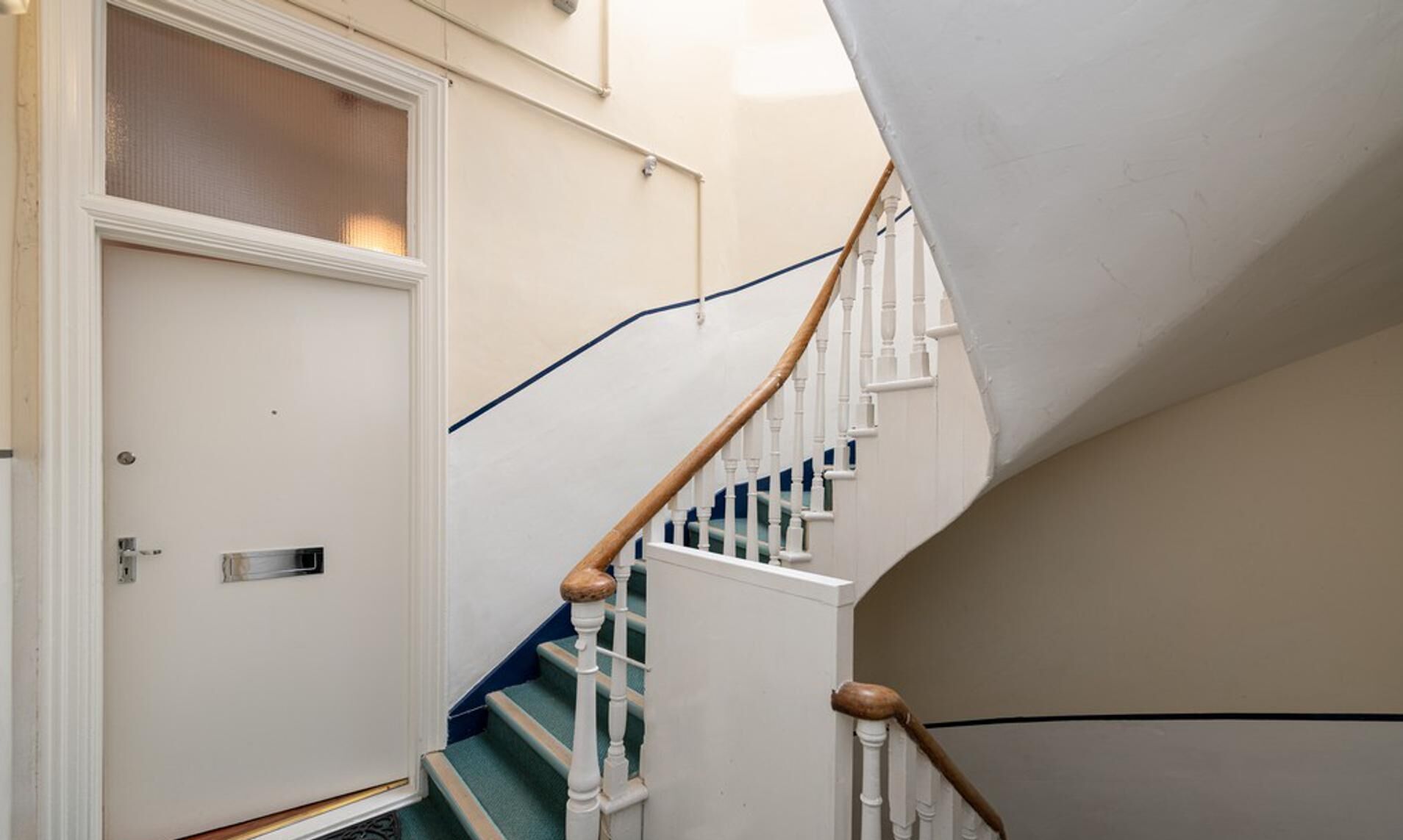 Un escalier avec des rampes blanches et des mains courantes en bois, menant à une porte dotée d’un panneau de verre.