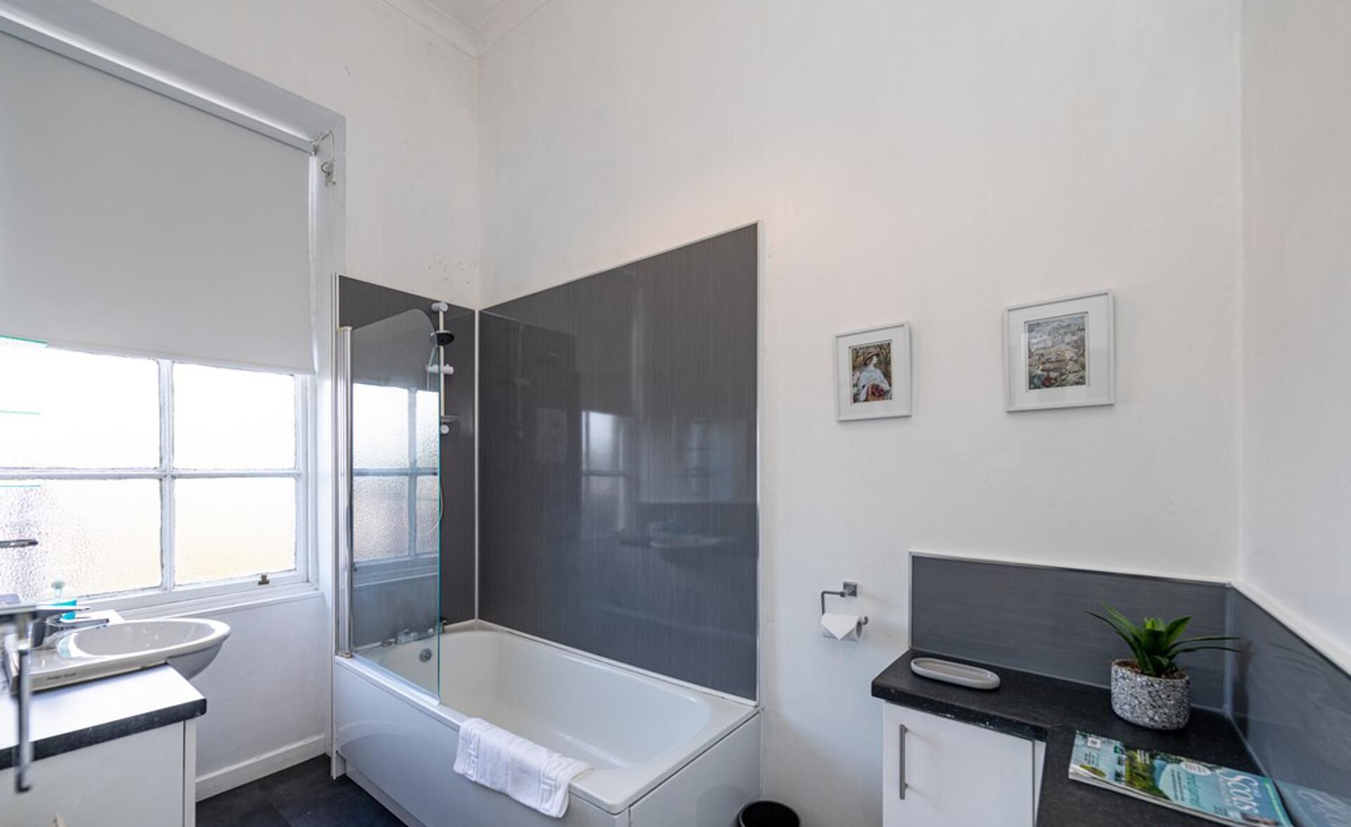A modern bathroom with a glass-enclosed shower, a white bathtub, a sink, and a small cabinet. There are two framed pictures on the wall.