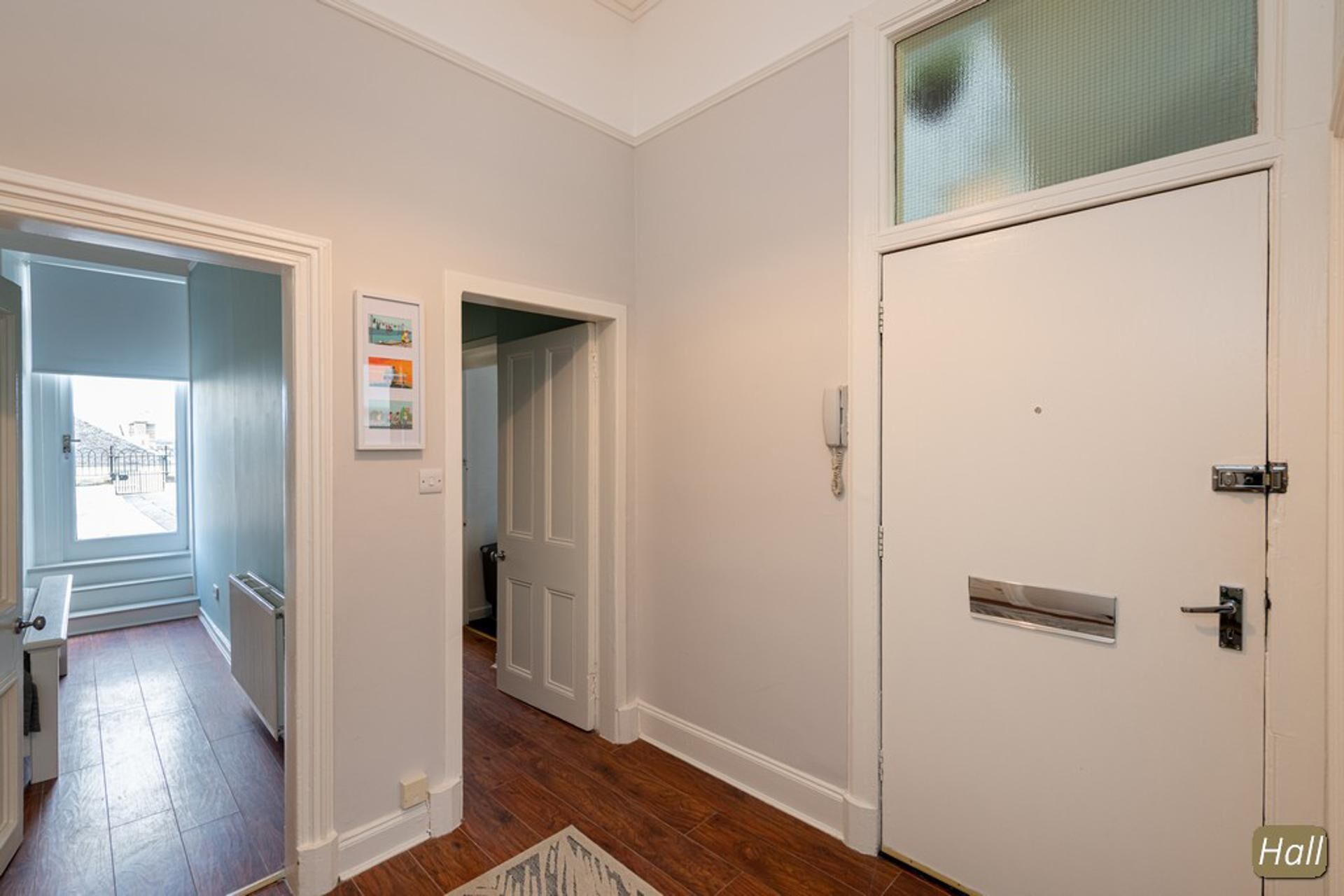 A hallway with a wooden floor, a white door with a silver handle, and a small picture frame on the wall.