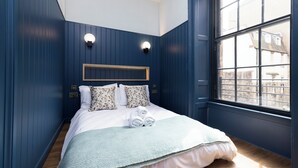 Quarto com uma janela grande, cama com roupa de cama branca e almofadas florais, e parede azul.