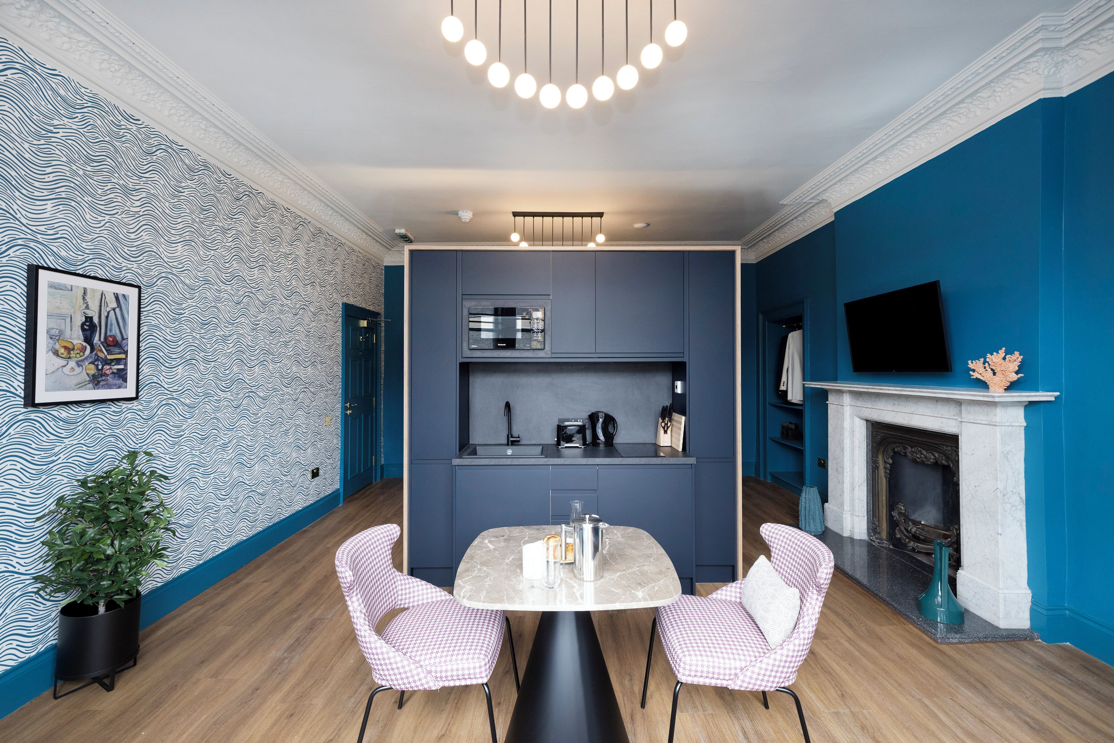 A modern kitchen with a built-in oven, microwave, and sink, a dining area with a marble table and checkered chairs, and a blue accent wall.