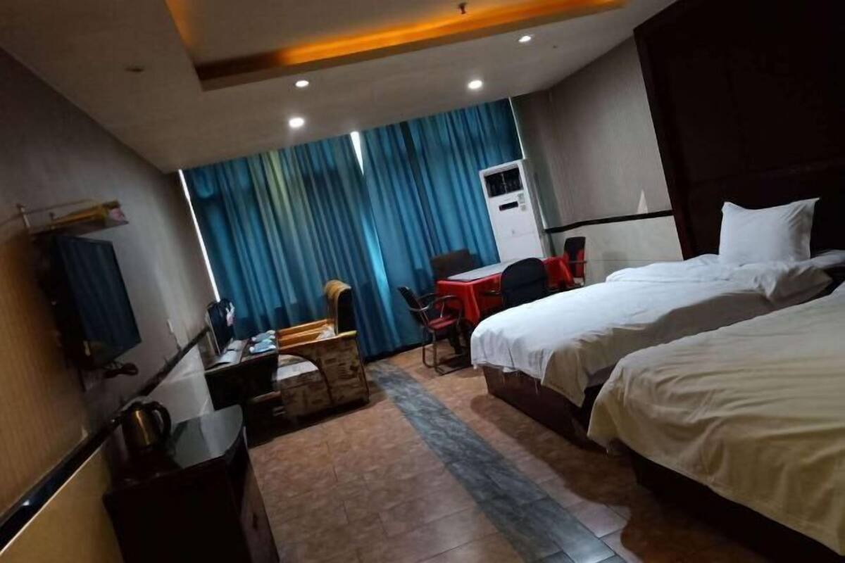 A hotel room with two beds, a desk, a chair, a small table, an air conditioning unit, and a curtain.